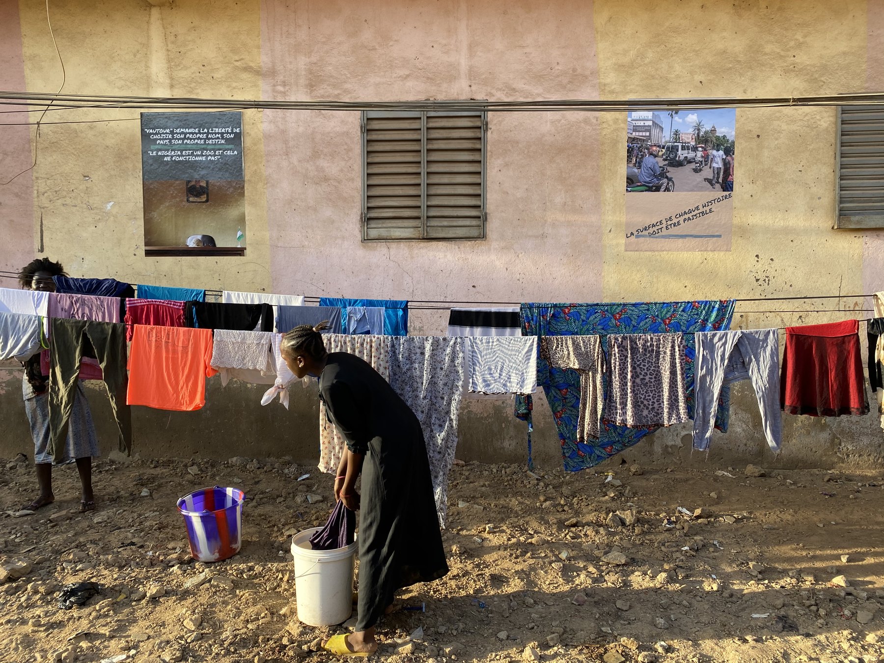 Two posters on a house wall above a full washing line, a woman bending over a bucket of laundry in the yard below and another at the far end of the line
