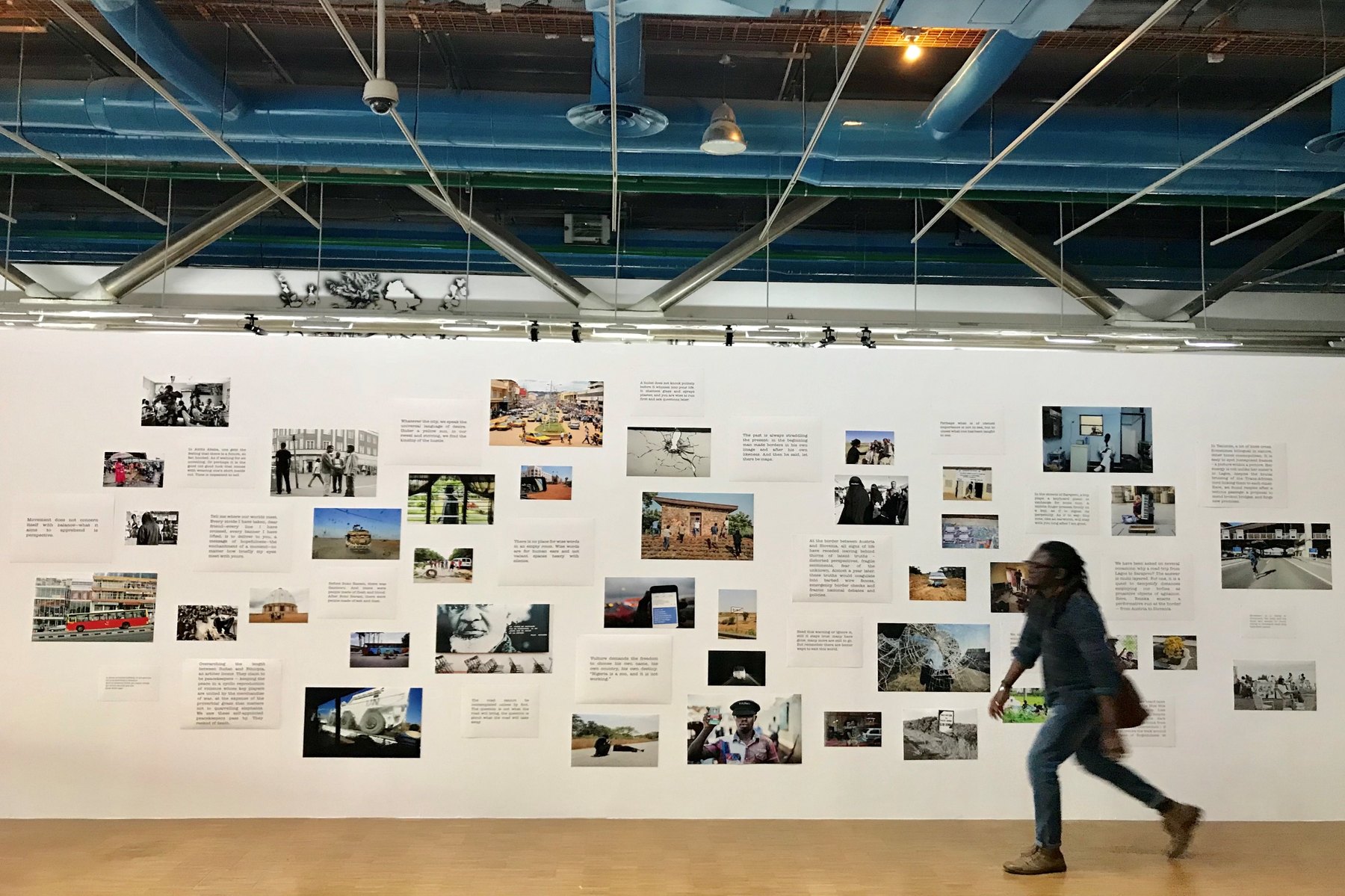 A long white wall carrying a constellation of small prints and text panels under exposed blue service ducts, a visitor walking past