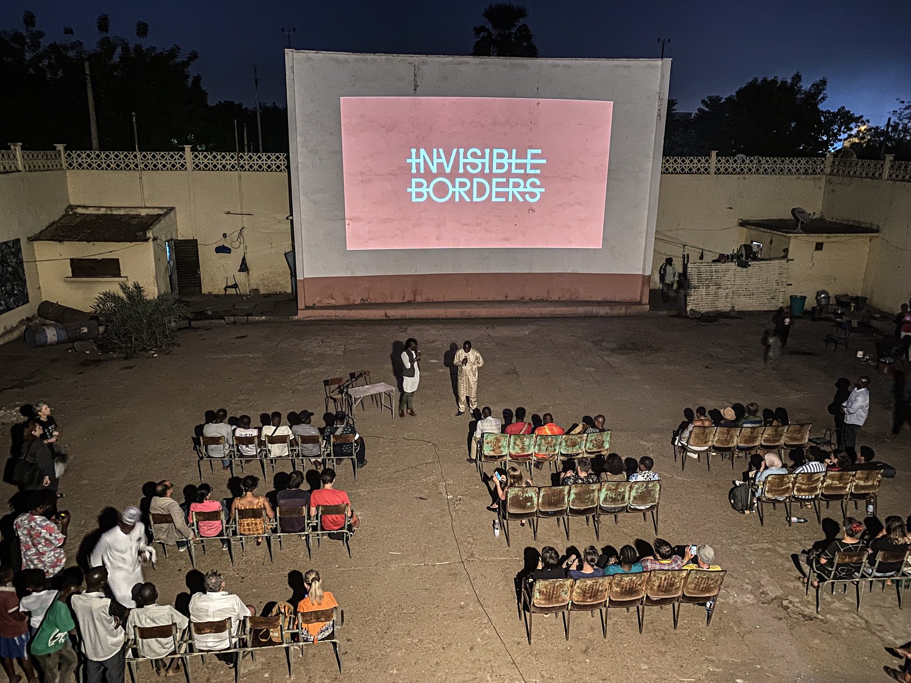 An audience seated on rows of chairs in a courtyard at dusk before a lit screen reading Invisible Borders