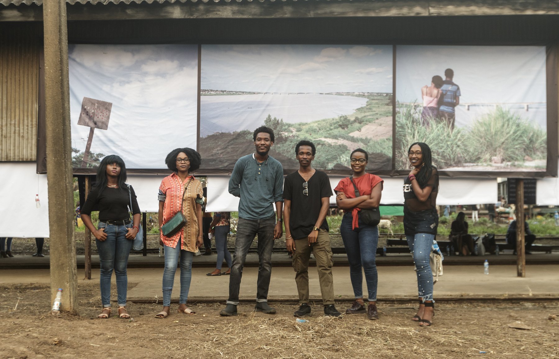 Seven young visitors standing on bare earth beneath large photographic banners strung under the railway shed roof, the open platform behind them