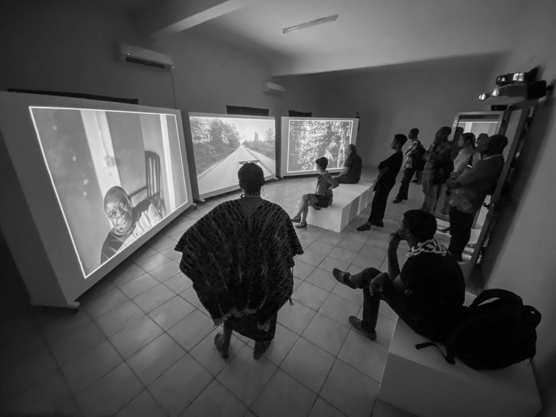 Large projection screens set in an L across a room, visitors seated on low benches, a standing figure in a dark boubou seen from behind