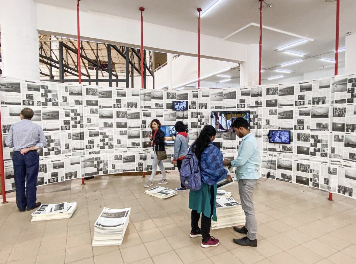 Freestanding walls papered edge to edge with newspaper pages and embedded monitors, stacks of the printed newspaper on the floor for visitors to take