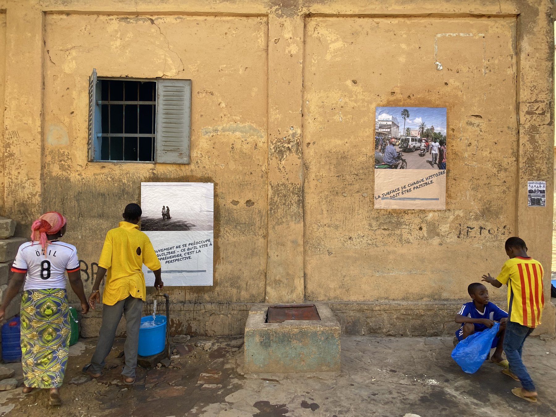 Two posters pasted on an ochre street wall beside a public standpipe, a woman and a man filling blue jerrycans, two boys passing at the right