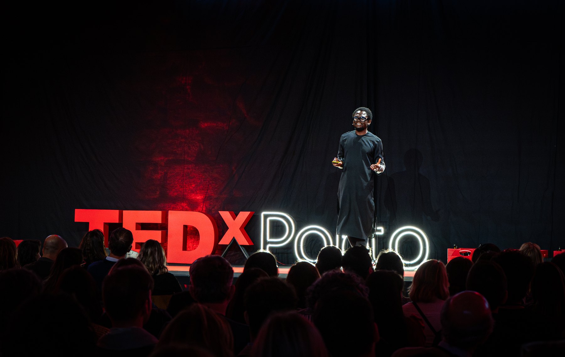 Emeka Okereke speaking on the TEDxPorto stage, the illuminated TEDxPorto lettering below him