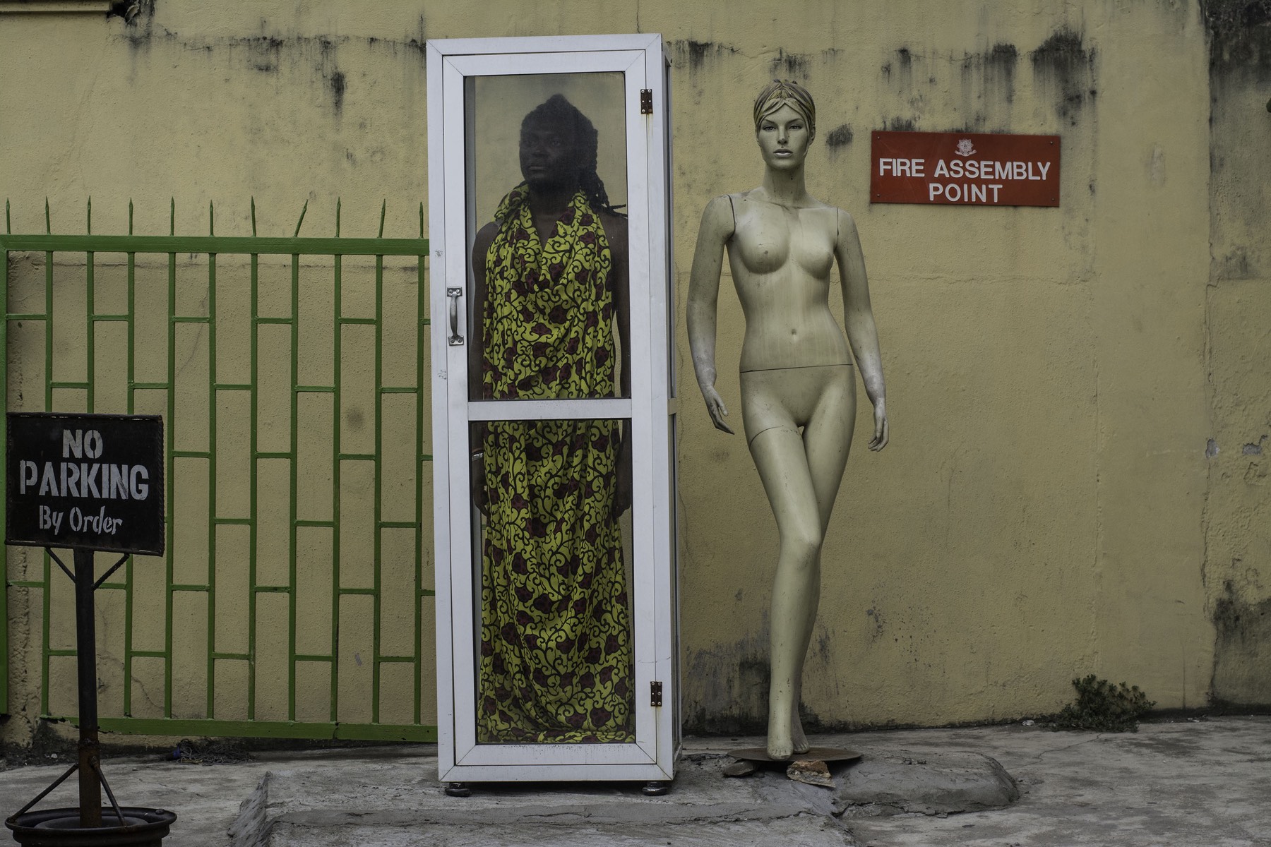 A figure behind a glass cabinet beside a mannequin against a weathered wall.