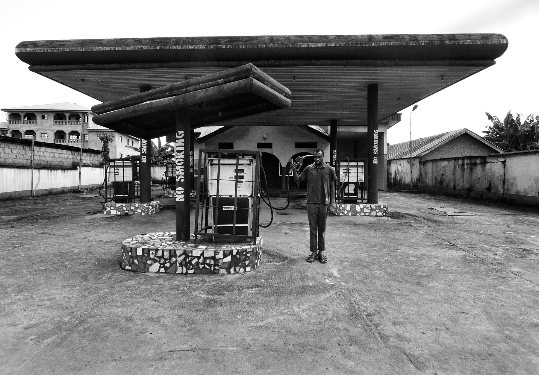 A man standing beneath the wide concrete canopy of a disused petrol station, holding a fuel nozzle to his head