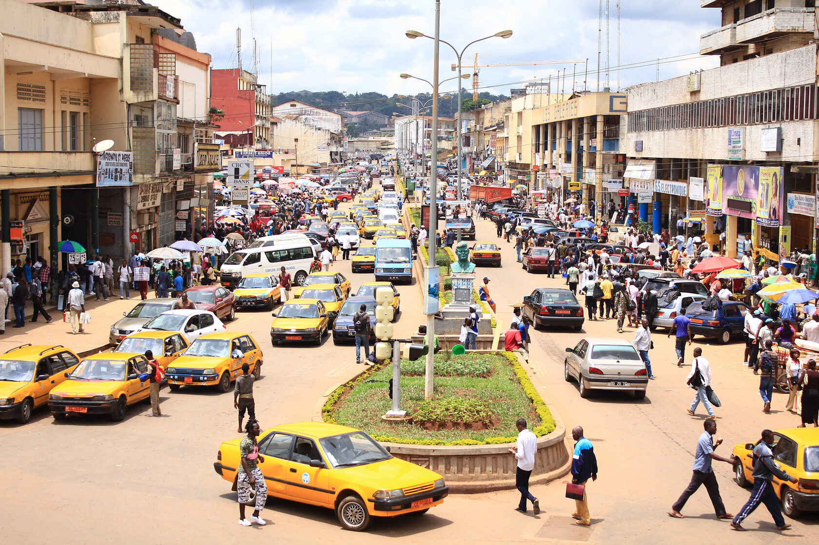 A busy street in Yaoundé seen from above: yellow taxis backed up at a junction, market umbrellas along the pavements and crowds crossing between them.